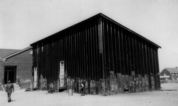 Black and white photograph of the temporary building constructed by Franconia Elementary School taken in 1954 for the Fairfax County School Board’s Fire Insurance Survey. The building was constructed out of wood and was covered with black tar-paper. Two entrance doors are visible. The building is an eye-sore, but was badly needed as the number of children living in the area far exceeded the number of classrooms available. 