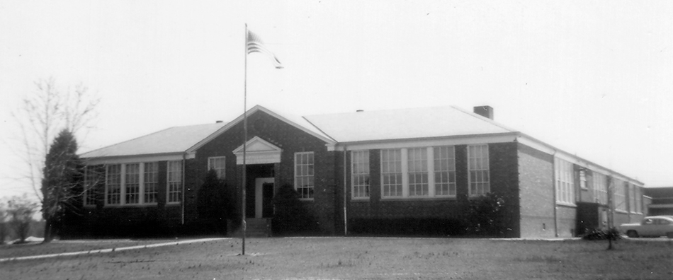 Black and white photograph of the original main entrance of Franconia Elementary School taken in 1954 for the Fairfax County School Board’s Fire Insurance Survey. A flagpole in visible in front of the building and several additions can be seen to the rear of the building on the far right of the image. Also on the far right is an early 1950s car. 