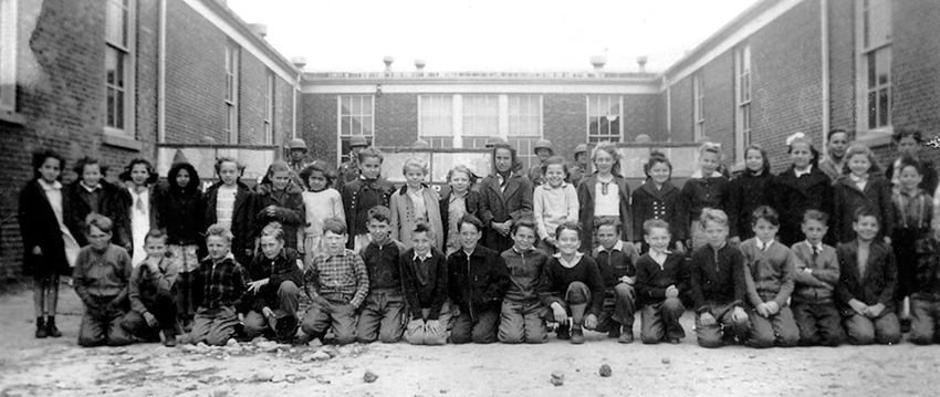 Black and white class photograph of children at Franconia Elementary School. The front row, all boys, are kneeling. Behind them stands a row of girls. The children are wearing coats and sweaters and look to be about 10-years-old. Behind them are U.S. Army soldiers sitting in jeeps. The picture was taken during the World War II timeframe, exact date unknown, but it appears to have been a visit to the school by soldiers and the photograph was taken to commemorate the occasion.  