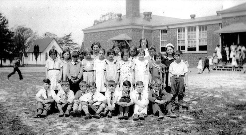 Black and white class photograph of a group of children with their teacher. The picture was taken outside the back of the original Franconia Elementary building as it appeared in 1932. A small church is visible in the distance. The building has had no additions constructed. Another group of children can be seen entering through a door in the rear of the building. The children in the photo appear about 10-years of age. The boys are seated on the ground and the girls and teacher stand behind them. The girls are wearing dresses and the boys are wearing short-sleeved shirts with ties so the picture was taken during warm weather.  