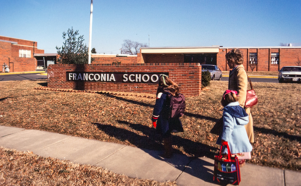 Color photograph from a 35 millimeter slide of the modern main entrance of Franconia Elementary School. The picture was taken in either the 1970s or 1980s. A mother and her two children are visible walking toward the school. 